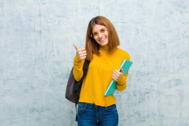 Genç bayan öğrenci neşeyle gülümsüyor ve mutlu görünüyor, kaygısız ve pozitif hissediyor, her iki başparmağı da grunge duvarının arkasında.