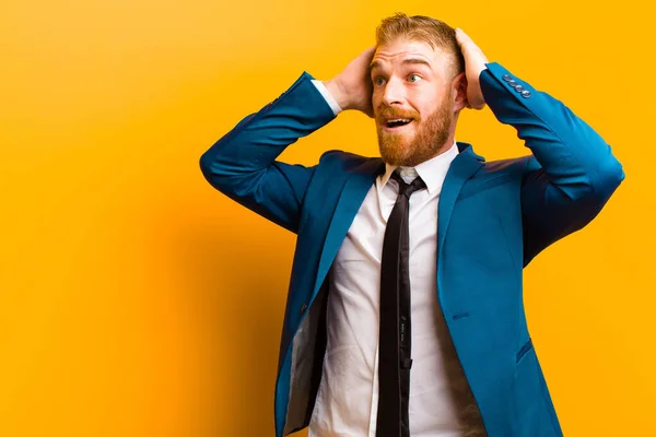 young red head businessman with open mouth, looking horrified and shocked because of a terrible mistake, raising hands to head against orange background