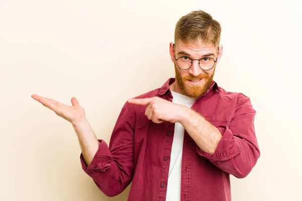 young red head man smiling cheerfully and pointing to copy space on palm on the side, showing or advertising an object against beige background