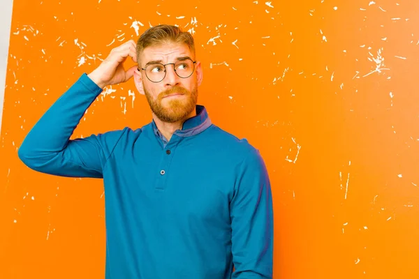 young red head man feeling puzzled and confused, scratching head and looking to the side against grunge orange wall