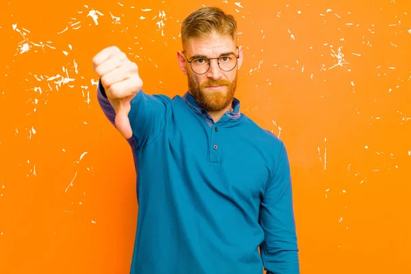 young red head man feeling cross, angry, annoyed, disappointed or displeased, showing thumbs down with a serious look against grunge orange wall