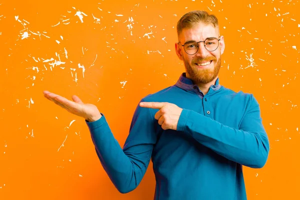 young red head man smiling, feeling happy, carefree and satisfied, pointing to concept or idea on copy space on the side against grunge orange wall