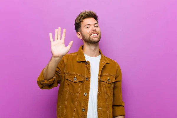 young man smiling happily and cheerfully, waving hand, welcoming and greeting you, or saying goodbye against purple background