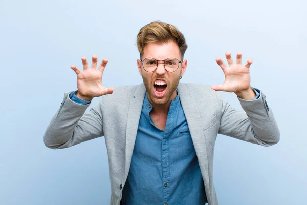 young businessman screaming in panic or anger, shocked, terrified or furious, with hands next to head against blue background