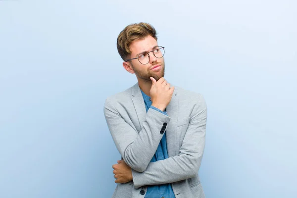 young businessman thinking, feeling doubtful and confused, with different options, wondering which decision to make against blue background