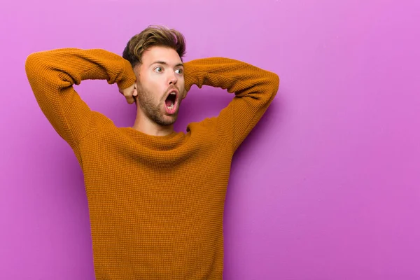 young man with open mouth, looking horrified and shocked because of a terrible mistake, raising hands to head against purple background