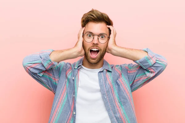 young man raising hands to head, open-mouthed, feeling extremely lucky, surprised, excited and happy against pink background