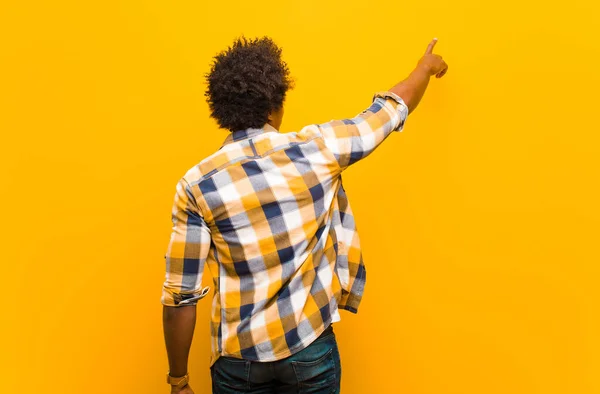 young black man standing and pointing to object on copy space, rear view against orange wall