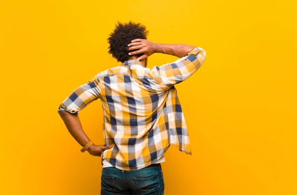 young black man thinking or doubting, scratching head, feeling puzzled and confused, back or rear view against orange wall