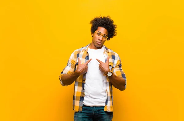 young black man pointing to self with a confused and quizzical look, shocked and surprised to be chosen against orange wall