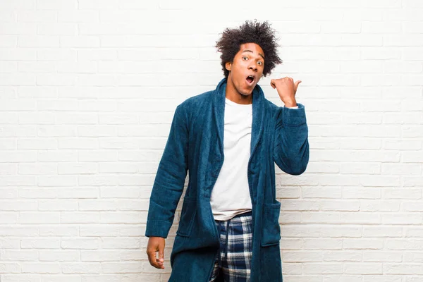 young black man wearing pajamas with gown looking astonished in disbelief, pointing at object on the side and saying wow, unbelievable against brick wall