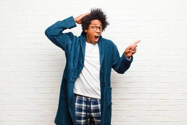 young black man wearing pajamas with gown laughing, looking happy, positive and surprised, realizing a great idea pointing to lateral copy space against brick wall