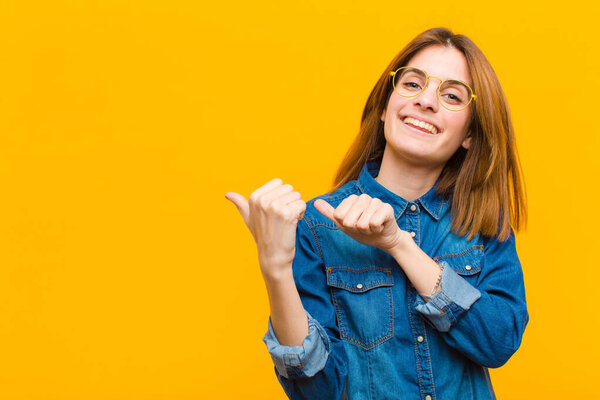 young pretty woman smiling cheerfully and casually pointing to copy space on the side, feeling happy and satisfied against yellow background