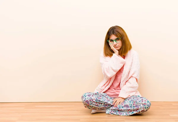 young woman wearing pajamas sitting at home feeling bored, frustrated and sleepy after a tiresome, dull and tedious task, holding face with hand