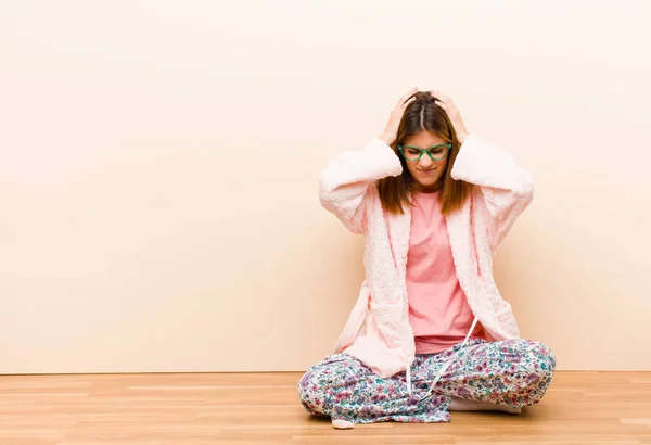 young woman wearing pajamas sitting at home feeling stressed and frustrated, raising hands to head, feeling tired, unhappy and with migraine