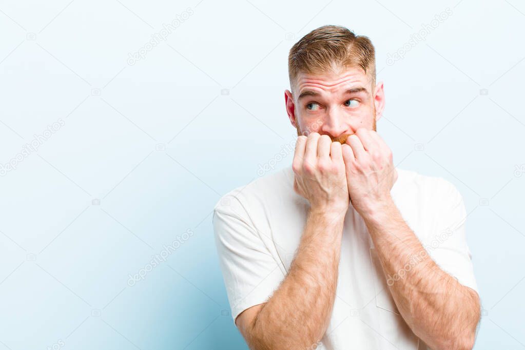 young red head man looking worried, anxious, stressed and afraid, biting fingernails and looking to lateral copy space against soft blue background
