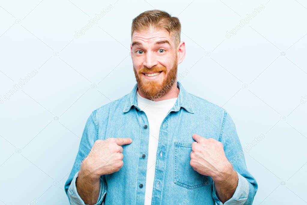 young red head man feeling happy, surprised and proud, pointing to self with an excited, amazed look against soft blue background