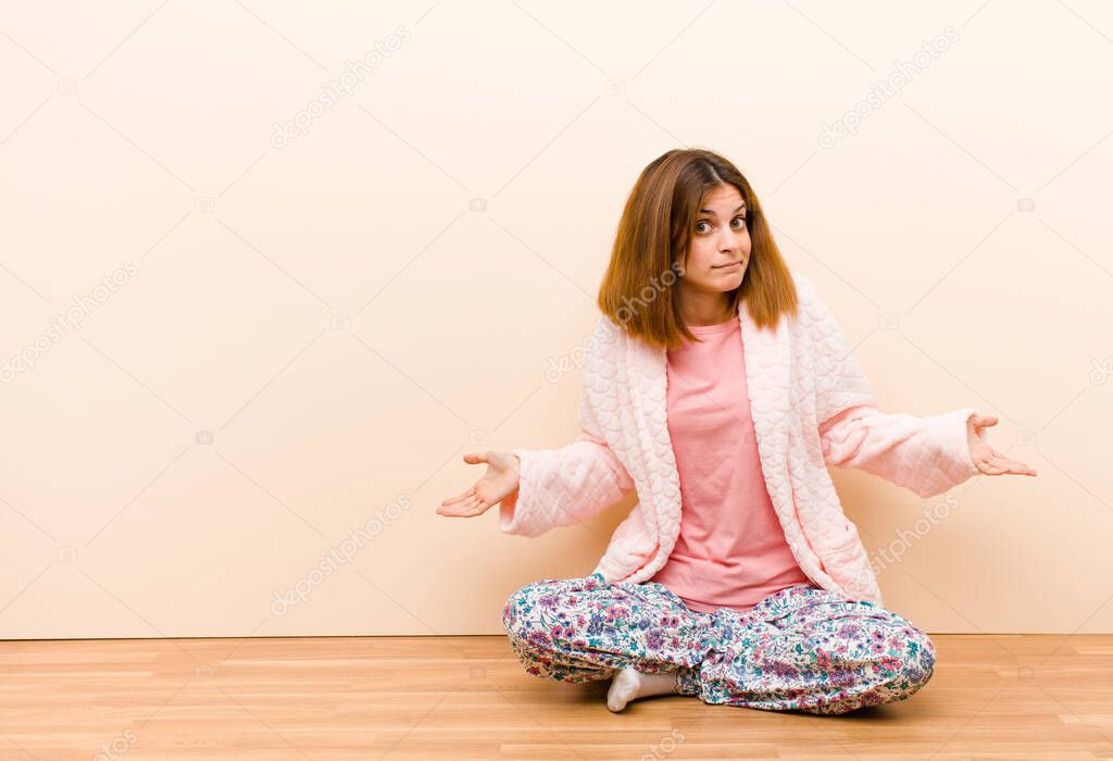 young woman wearing pajamas sitting at home feeling clueless and confused, having no idea, absolutely puzzled with a dumb or foolish look