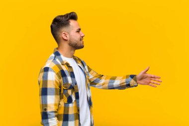 Genç İspanyol adam gülümsüyor, seni selamlıyor ve başarılı bir anlaşma için el sıkışmayı teklif ediyor. Turuncu duvara karşı işbirliği.