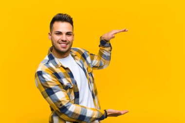 Genç İspanyol adam gülümsüyor, mutlu, pozitif ve tatmin olmuş hissediyor, fotokopi odasında turuncu duvara karşı nesne ya da konsept tutuyor ya da gösteriyor