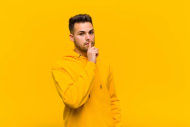 Genç İspanyol adam sessizlik ve sessizlik istiyor, parmaklarıyla ağzının önünde işaret ediyor, ya da turuncu duvara karşı bir sır saklıyor.