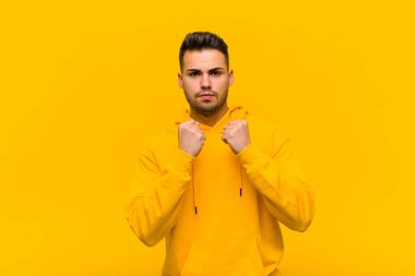 Genç İspanyol adam kendinden emin, sinirli, güçlü ve agresif görünüyor. Turuncu duvara karşı boks pozisyonunda dövüşmeye hazır yumrukları var.