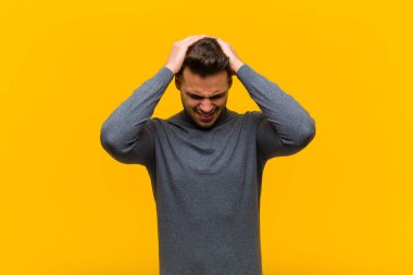 Genç İspanyol adam stresli ve hayal kırıklığına uğramış hissediyor. Yumruk yumruğa, yorgun, mutsuz ve migreni turuncu duvara dayanmış.