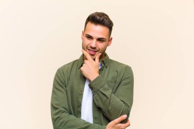 Genç İspanyol bir adam mutlu ve kendinden emin bir ifadeyle gülümsüyor. Çenesinde elleri olan, izole edilmiş arka plana bakıp merak eden ve yan tarafa bakan.