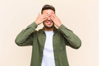 Genç İspanyol adam gülümsüyor ve mutlu hissediyor, iki eliyle gözlerini kapatıyor ve izole edilmiş arka planda inanılmaz bir sürpriz bekliyor.