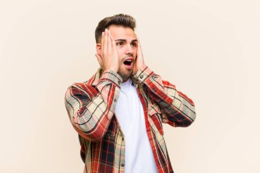 Genç İspanyol bir adam mutlu, heyecanlı ve şaşırmış hissediyor. İki eli yüzü üzerinde, izole edilmiş arka plana bakıyor.