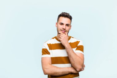 Genç İspanyol adam mutlu görünüyor ve gülümsüyor, çenesi açık, merak ediyor ya da soru soruyor, mavi duvarla seçenekleri karşılaştırıyor.