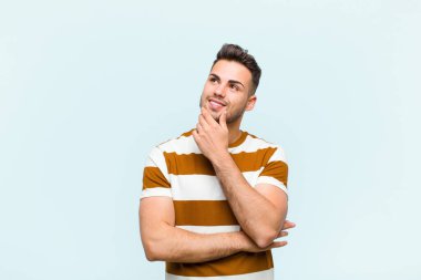 Genç İspanyol bir adam mutlu, kendinden emin bir ifadeyle gülümsüyor. Çenesinde elleri, mavi duvara bakıyor ve merak ediyor.