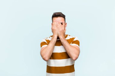 Genç İspanyol adam elleriyle yüzünü kapatıyor, parmaklarının arasından şaşkın bir ifadeyle bakıyor ve mavi duvara bakıyor.