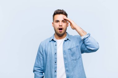 Genç İspanyol adam mutlu, şaşkın ve şaşırmış görünüyor, gülümsüyor ve mavi duvara karşı inanılmaz ve inanılmaz iyi haberler veriyor.