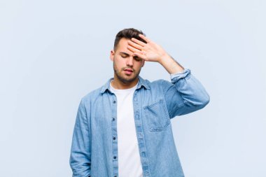 Genç İspanyol adam stresli, yorgun ve hayal kırıklığına uğramış görünüyor, alnından ter akıyor, mavi duvara karşı umutsuz ve bitkin hissediyor.