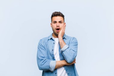 Genç İspanyol adam şok ve inançsızlıkla ağzı açık, elleri yanak ve kolları çaprazlanmış, sersemlemiş ve mavi duvara karşı şaşkın hissediyordu.