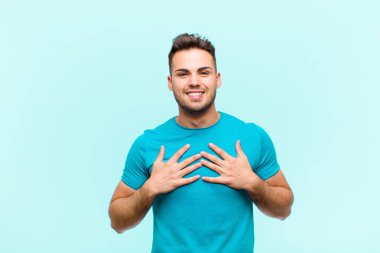 Genç İspanyol adam mutlu, şaşırmış, gururlu ve heyecanlı görünüyor. Mavi arka planda kendini işaret ediyor.