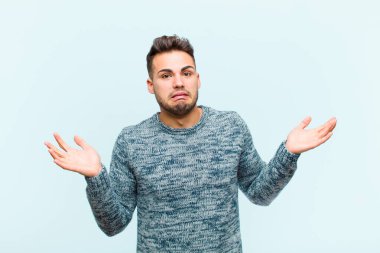 Genç İspanyol adam aptal, deli, kafası karışık, şaşkın bir ifadeyle omuz silkiyor. Mavi arka plana karşı sinirli ve bilgisiz hissediyor.
