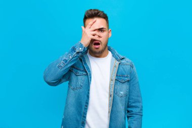 Genç İspanyol adam şok olmuş, korkmuş ya da korkmuş görünüyor. Yüzü elle kaplanmış ve parmaklarının arasından mavi arka plana bakıyor.