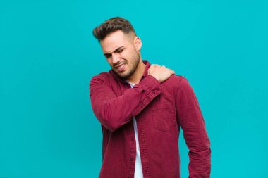 Genç İspanyol adam yorgun, stresli, endişeli, hayal kırıklığına uğramış ve depresif hissediyor. Mavi arka planda sırt ya da boyun ağrısı çekiyor.