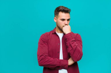 Genç İspanyol bir adam ciddi, düşünceli ve endişeli, elleri çenesine değmiş mavi arka planda yan bakıyor.