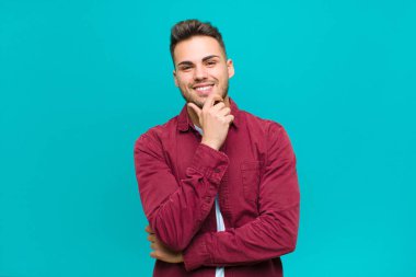 Genç İspanyol adam gülümsüyor, hayattan zevk alıyor, mutlu, arkadaş canlısı, tatmin olmuş ve elleri mavi arka planda tasasız.