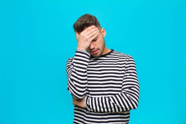Genç İspanyol bir adam stresli, utanmış ya da üzgün görünüyor, baş ağrısı var, yüzünü mavi arka planda kapatıyor.