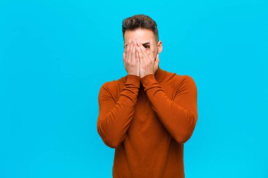 Latin kökenli genç bir adam korkmuş ya da utanmış hissediyor. Gözleri yarı kapalı mavi arka planda dikizliyor ya da dikizliyor.