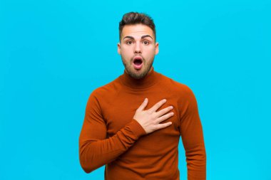 Genç İspanyol adam şok olmuş ve şaşırmış hissediyor, gülümsüyor, el ele tutuşuyor, mutlu ya da mavi arka plana karşı minnettarlık gösteriyor.