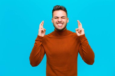 Genç İspanyol adam gergin ve umutlu, dua ediyor ve mavi arka planda iyi şanslar diliyor.