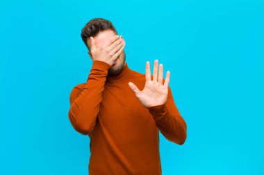 Latin asıllı genç bir adam yüzünü eliyle kapatıyor ve diğer elini kamerayı durdurmak için öne koyuyor. Mavi arka plandaki fotoğraf ve resimleri reddediyor.