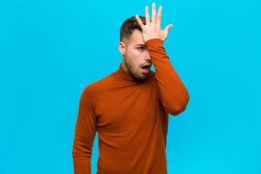 Genç İspanyol bir adam, aptalca bir hata yaptıktan ya da hatırladıktan sonra mavi arka planda kendini aptal gibi hissederek avuçtan avuca kadar yükseltiyor.