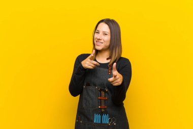 Pozitif, başarılı, mutlu bir tavırla gülümseyen kuaför kadın kamerayı işaret ederek, elleri turuncu arka planda silah işareti yapıyor.