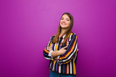 Genç ve güzel bir kadın kollarını kavuşturup mutlu bir şekilde gülüyor. Mor arka planda rahat, pozitif ve tatmin edici bir pozla.